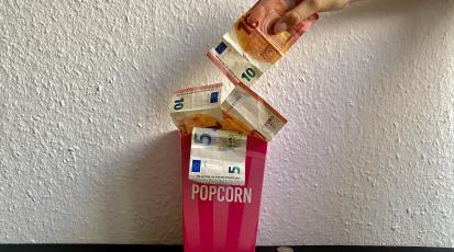 Eine Schachtel mit Popcorn, aus der Geld herauskommt, und eine Hand nimmt einen Geldschein aus der Schachtel.