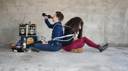 Zwei Personen sitzen auf dem Boden. Die beiden sind durch ein Seil aneinander gebunden. Die linke Person trinkt aus einer Weinflasche, neben ihr stehen Bierkästen und Alkoholflaschen. Die rechte Person hat ihr Gesicht in ihren Händen versteckt.