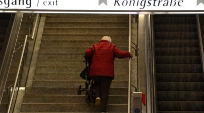 Ältere Dame mit rotem Mantel trägt ihre Gehhilfe die Treppen von einer U-Bahn Station hoch.