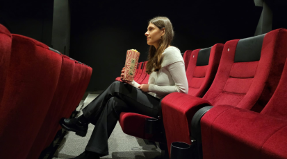 Eine Frau sitzt mit überkreuzten Beinen auf einem roten Kinosessel und schaut gespannt auf die Leinwand. Sie hält eine Popcorntüte in der Hand.