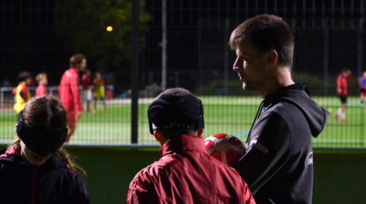 Alex redet mit zwei Mitspielern beim Training, im Hintergrund sieht man andere sehende Jugendliche Fußball spielen.