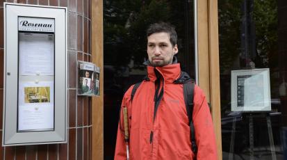Florian Günther steht vor dem Eingang des Restaurants Rosenau in Stuttgart, das drei Tage im Monat zu einem Dunkelrestaurant umfunktioniert wird. Florian trägt eine rote Jacke. In der rechten Hand hält er seinen Blindenstock nah an seinem Körper. 
