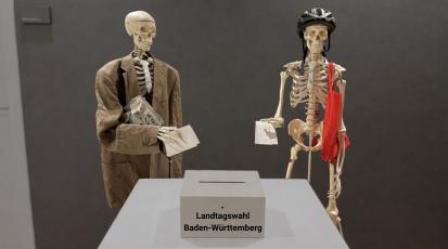 Zwei Skelette, die ihre Wahlzettel in eine Urne der Landtagswahl Baden-Württemberg schmeißen
