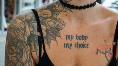 Auf dem Foto ist das Dekoltee ener Frau. Im Mittepunkt ist ein Tattoo zu sehen in schwarzer Schrift. Es steht dort "my body my choice".