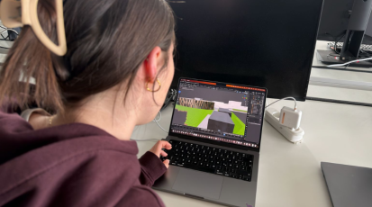 Studentin sitzt links vor ihrem Laptop auf dem ein 3D-Model der Hochschule der Medien zu sehen ist.