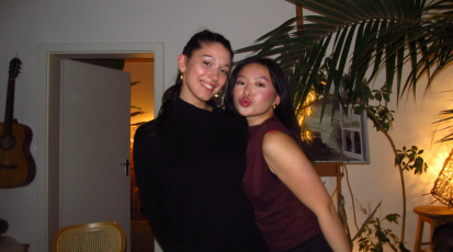 Zwei junge Frauen stehen eng nebeneinander in einem Wohnzimmer und lächeln in die Kamera. Im Hintergrund sind Pflanzen, eine offene Tür und warmes Licht zu sehen.