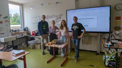 Die Studierenden und ein Grundschullehrer stehen gemeinsam vor einer digitalen Tafel, auf der die WebApp zu sehen ist.