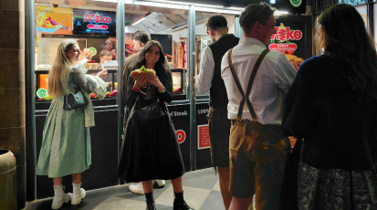 In der Mitte des Bilds ist eine Frau in einem Dirndl vor einem Dönerladen zu sehen, die einen Döner in ihren Händen hält. Auf der rechten Seite des Bilds sieht man eine Warteschlange. Auf der linken Seite nimmt gerade eine Frau einen Döner entgegen. Alles Personen auf dem Bild sind mit Dirndl oder Lederhose gekleidet. 