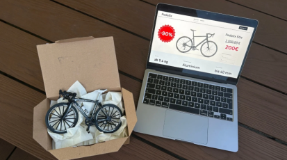 Rechts auf dem Bild ist ein Laptop auf welchem ein Rennrad online günstig Angeboten wird. Auf der linken Seite im Bild ist ein Karton in welchem ein Miniaturrennrad zu sehen ist.