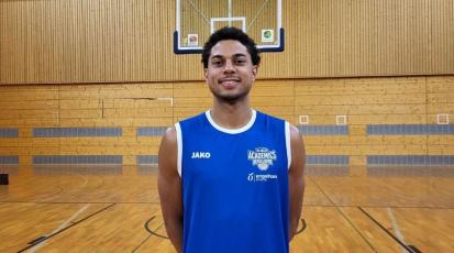 Auf dem Bild ist ein Mann in einem blauen Basketballtrikot zu sehen. Er schaut frontal in die Kamera. Im Hintergrund ist ein Basketabllkorb zu sehen.