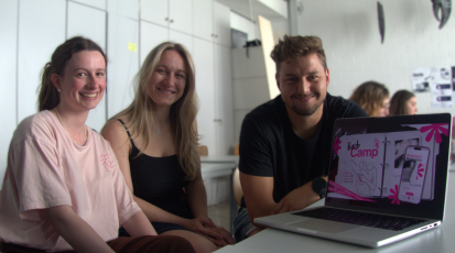 Drei Mitglieder der "Boob-Camp"-Gruppe lächeln neben einem offenen Laptop, auf dem ihr Projekt zu sehen ist, in die Kamera.