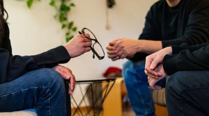 Frau, die Brille in der Hand hält und gegenüber einem Pärchen sitzt.