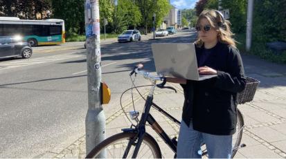 Eine Frau steht an einer Ampel. In einer Hand hält sie einen Laptop, in der anderen ein Fahrrad. 