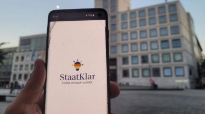 Bild eines Handys vor dem Stuttgarter Rathaus. Auf dem Handy ist das "StaatKlar"-Logo zu sehen.