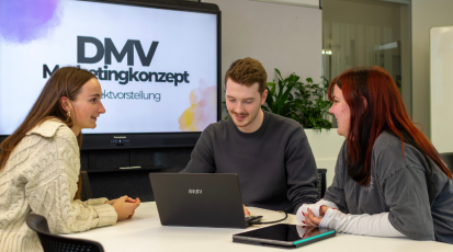 Studierende der HdM sitzen an einem Tisch, diskutieren angeregt und planen ein Marketing-Projekt; im Hintergrund ein Bildschirm mit „DMV-Marketing-Konzept“.