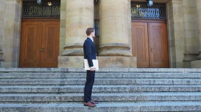 Ein Mann im Anzug/Business Kleidung steht auf einer Treppe und dreht seinen Kopf unentschlossen in Richtung von zwei Türen. Er überlegt durch welche der beiden er gehen soll.