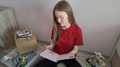 Mädchen sitzt auf dem Boden und blättert ein Buch durch. Um sie herum Kisten voll mit Kindheitserinnerungen.