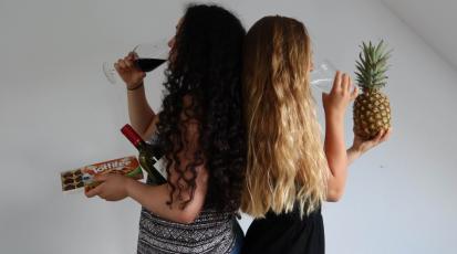 Zwei Frauen stehen Rücken an Rücken. Während eine der Frauen Wein trinkt und Schokolade in der Hand hält, trinkt die andere Wasser und hält eine Ananas in der Hand.
