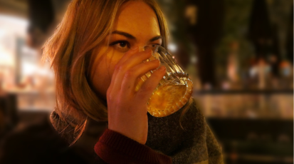 Eine Frau, die draußen in einer Bar sitzt und ein Glas mit Whiskey zum trinken ansetzt.