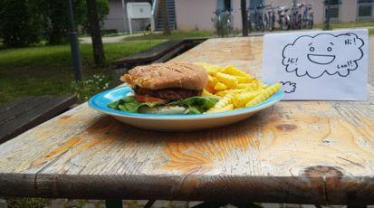 Auf einem blauen Teller liegt ein Burger mit Pommes. Daneben ist ein Blatt Papier aufgestellt, auf dem eine Gedankenwolke mit einem lachenden Gesicht und verschiedenen Worten gezeichnet. Alles steht auf einem Biertisch in einem Innenhof.