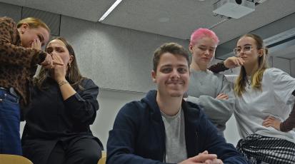 Ein Mann schaut lächelt künstlich in die Kamera. Im Hintergrund blickt eine Gruppe auf ihn.