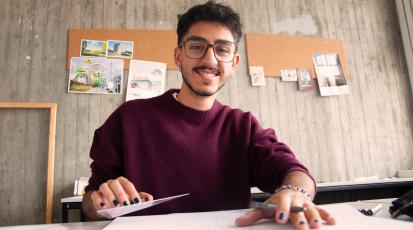 Ein junger Mann sitzt zeichnend am Tisch und blickt lächelnd in die Kamera. 