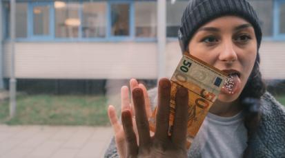 Junge Frau mit Globuli auf der Zunge drückt 50 Euro Schein an Fensterscheibe.