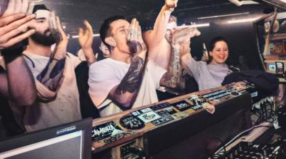 Feiernde, jubelnde Leute stehen vor einem DJ Pult