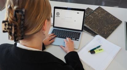 Eine junge Frau in Arbeitskleidung sitzt an einem Tisch. Auf dem Tisch liegen Arbeitsmaterialien wie Ordner und ein Block mit Stiften. Die Frau ist jedoch konzentriert auf ihren Laptop, der vor ihr steht, auf dem sie ChatGPT geöffnet hat.