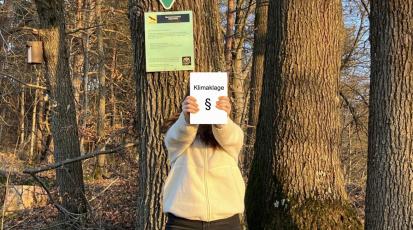 Person steht im Wald und hält ein Buch auf dem „Klimaklage“ steht