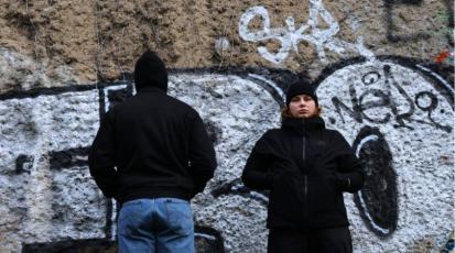 Zwei Menschen stehen vor einer Mauer, einer mit dem Rücken zur Mauer, der andere sieht die Mauer an.