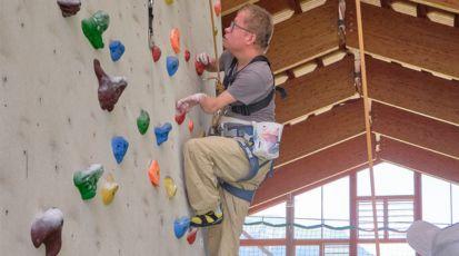 Marcel Richter klettert an einer Kletterwand hoch. Er ist kleinwüchsig und deformierte Hände. Er trägt eine Klettermontur, in Hintergrund ist ein Panoramafenster zu sehen.