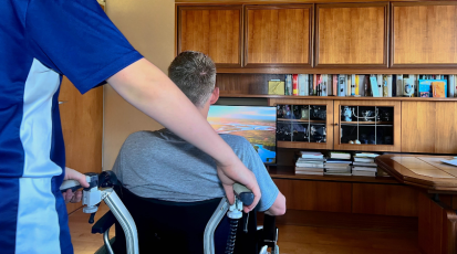 Ein Kind steht hinter seinem Vater, der im Rollstuhl vor dem Fernseher sitzt.