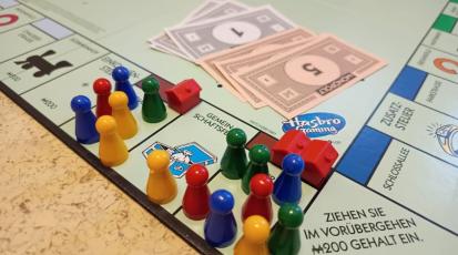 Zu sehen ist ein Ausschnitt eines Monopoly Spielbretts. Man sieht zahlreiche Spielfiguren, die um wenig verfügbaren Wohnraum konkurrieren. 