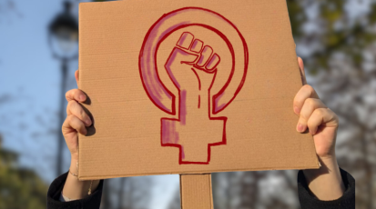 Pappschild mit feministischem Symbol: Erhobene Faust in einem Venuszeichen, gehalten von Händen vor unscharfem Hintergrund. Symbol für den Kampf um Gleichberechtigung und Sichtbarkeit von Frauen.