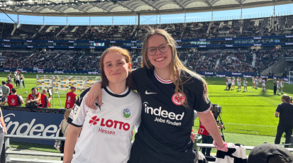 Zwei Fans lächeln und stehen Arm in Arm auf der Tribüne des Deutschen Bank Parks. Eine trägt ein Trikot des 1. FFC Frankfurt, die andere trägt eines mit dem Adler der Eintracht Frankfurt, im Hintergrund sieht man den Deutsche Bank Park.