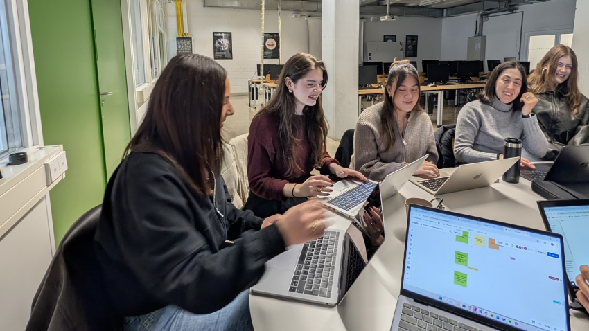 Fünf junge Frauen sitzen an einem Tisch und schauen in ihre Laptops.