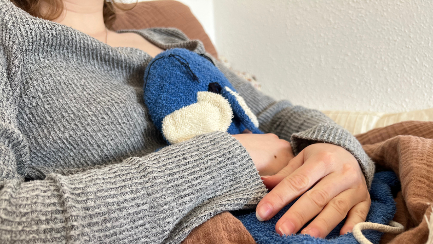 Zu sehen ist ein Ausschnitt, bei dem ein Mädchen eine Wärmflasche gegen ihren Bauch drückt.