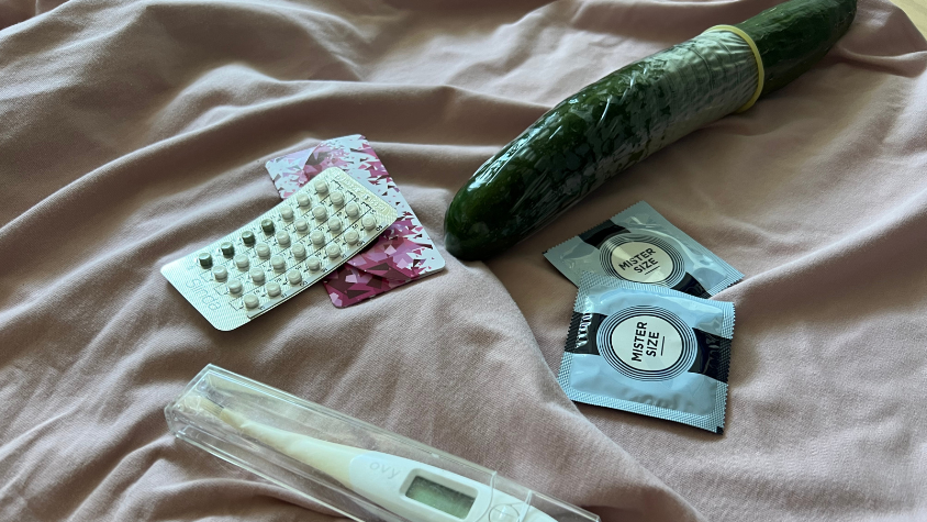 Zu sehen sind die Minipille, ein Basalthermometer, zwei Kondomverpackungen und eine Gurke die mit einem Kondom überzogen ist.