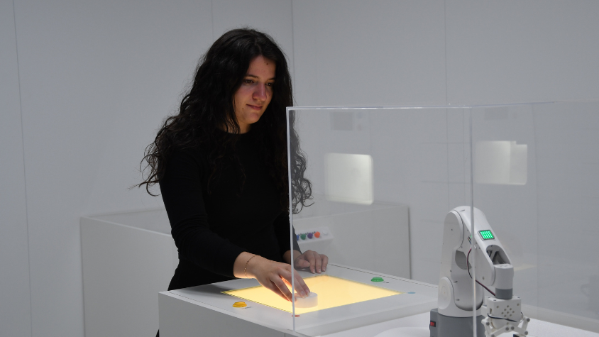 Frau steht an einer Station, an der sie ein Robotikarm steuern kann.