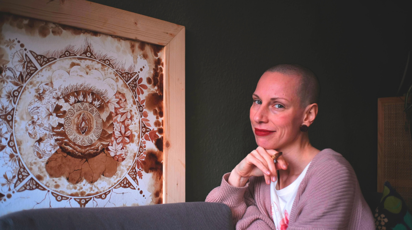 Maria sitzt vor einem selbst gemalten Gemälde, der Hintergrund ist schwarz. Sie hält einen Pinsel in ihrer Hand.