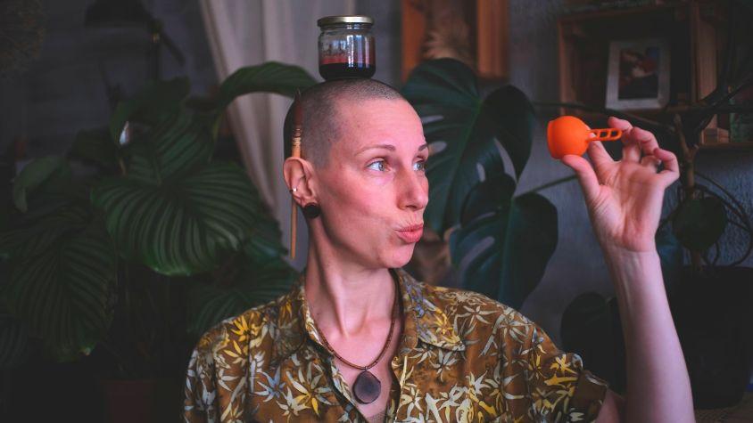 Maria hat ein Glas mit Menstruationsblut auf dem Kopf und hält ihre Menstruationstasse hoch.
