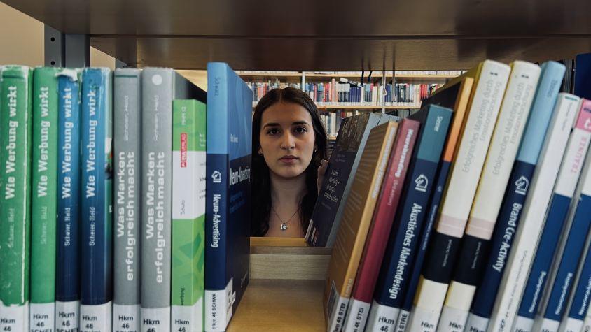 Ein Mädchen steht vor der Lücke eines Bücherregals. Ihr Gesichtsausdruck ist ernst.