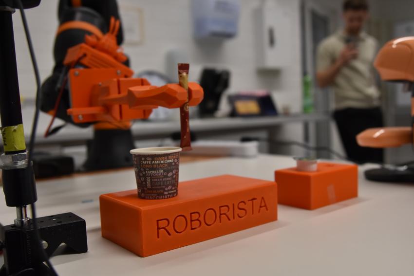 Der orangene Arm des Kaffeeroboters serviert ein Tütchen Zucker zum Espresso. 