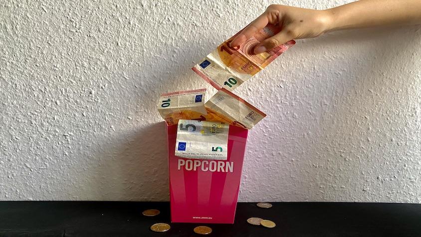 Eine Schachtel mit Popcorn, aus der Geld herauskommt, und eine Hand nimmt einen Geldschein aus der Schachtel.