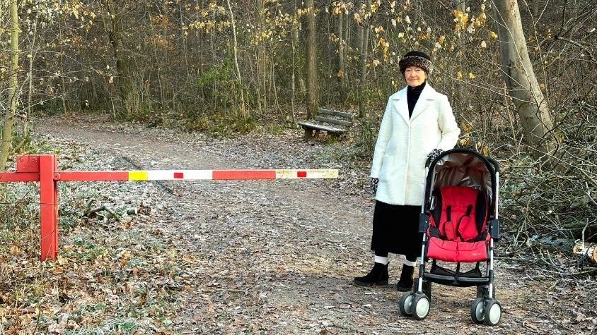Eine ältere Frau steht neben einem leeren Kinderwagen in einer Waldlichtung direkt vor einer Schranke.