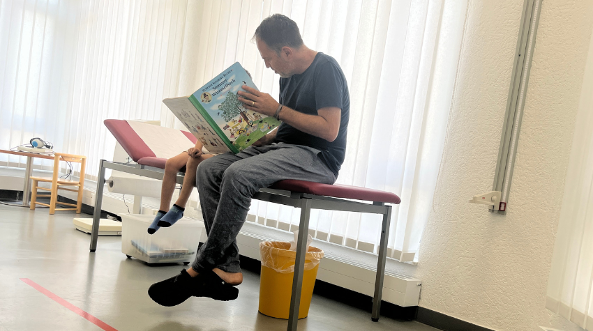 Der Kinderarzt sitzt auf einer Arztliege und zeigt einem kleinen Jungen ein Buch.