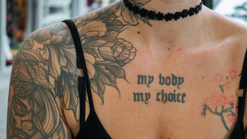 Auf dem Foto ist das Dekoltee ener Frau. Im Mittepunkt ist ein Tattoo zu sehen in schwarzer Schrift. Es steht dort "my body my choice".