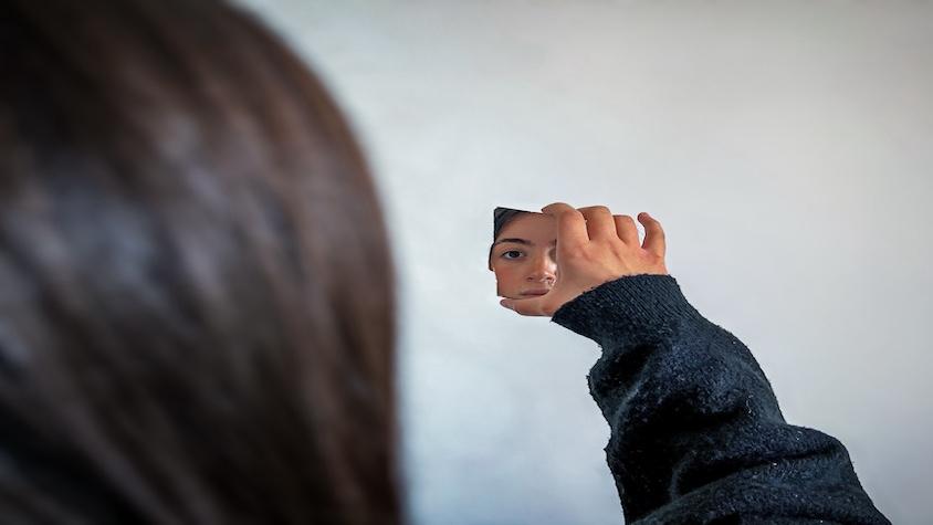 Frau mit der Scherbe eines zerbrochenen Spiegels in der Hand, in der sich ein Teil ihres Gesichts spiegelt.