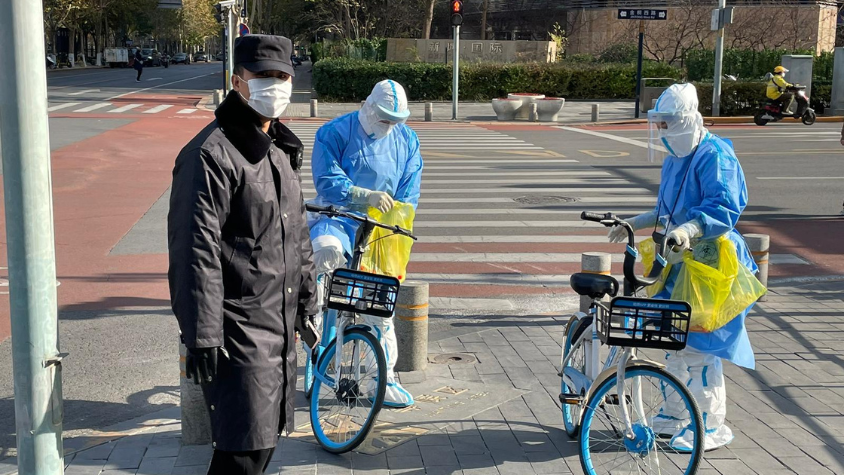 Ein Einwohner Pekings mit Mund-Nasen-Schutz, bei ihm zwei Angestellte einer Teststelle in Corona Schutzkleidung, die auf Fahrrädern unterwegs sind. 